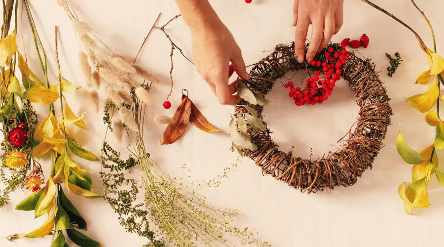 Zwei Hände einer Frau dekorieren einen Kranz mit herbstlich gefärbten Pflanzen, einschließlich roter Vogelbeeren, auf einer weißen Unterfläche.