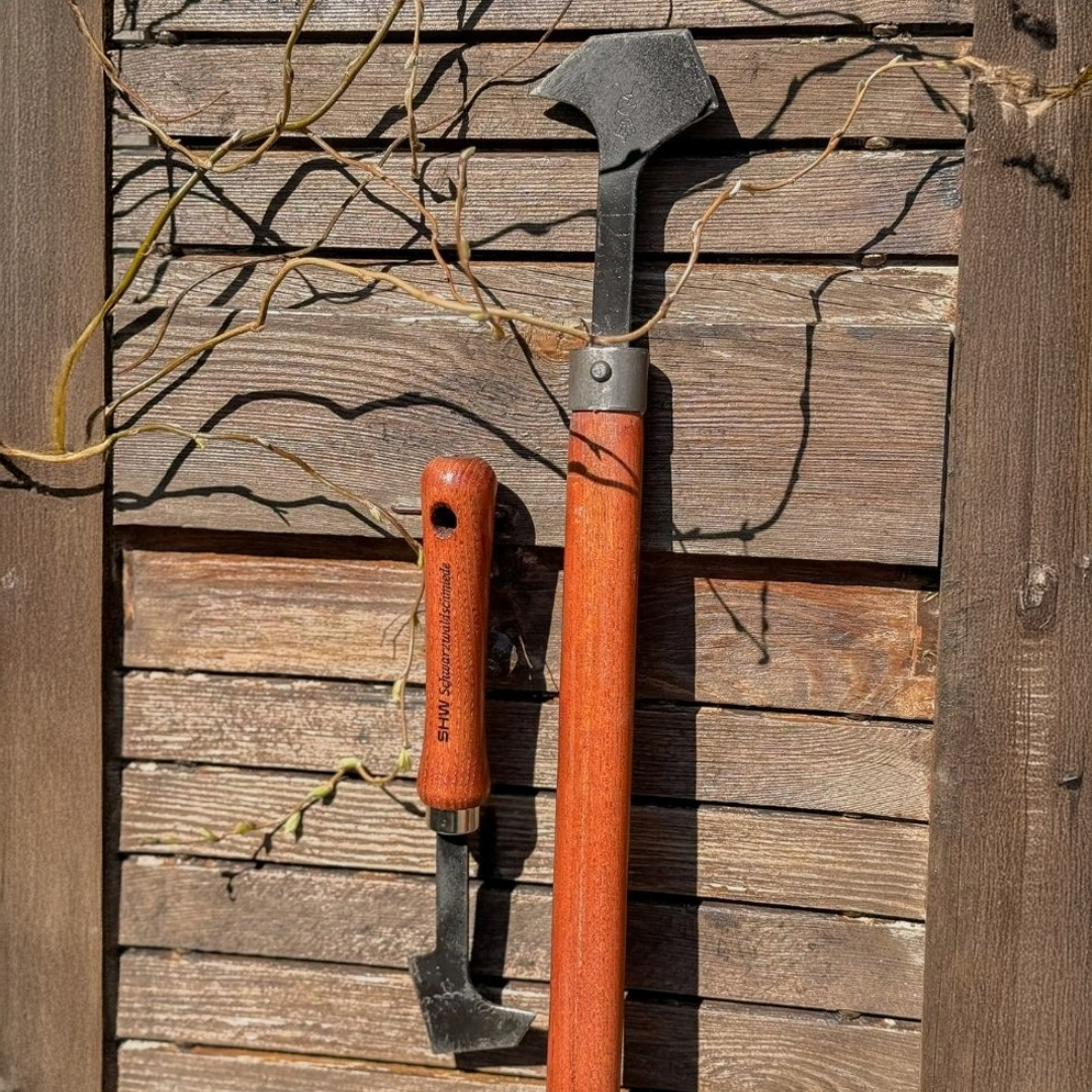 Zwei SHW Fugenkratzer mit kurzen und langen Holzgriffen und abgewinkelten Stahlklingen vor einer Holzwand. Gartenwerkzeuge zum Entfernen von Unkraut aus Pflasterfugen.