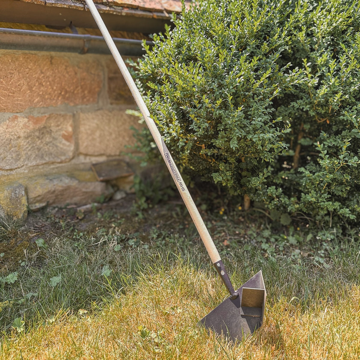 SHW Rasenkantenschneider mit langem Holzstiel und geschmiedetem Stahlblatt auf einer Rasenfläche vor einer Hecke. Gartenwerkzeug zum präzisen Abstechen von Rasenkanten und Formen sauberer Beetkanten.