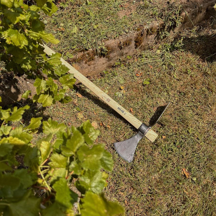 SHW Wiedehopfhacke mit langem Holzstiel, breitem Hackenblatt und gegenüberliegender Spitze auf einer Rasenfläche neben einem Gartenbeet. Zweischneidiges Gartenwerkzeug für feste Böden.
