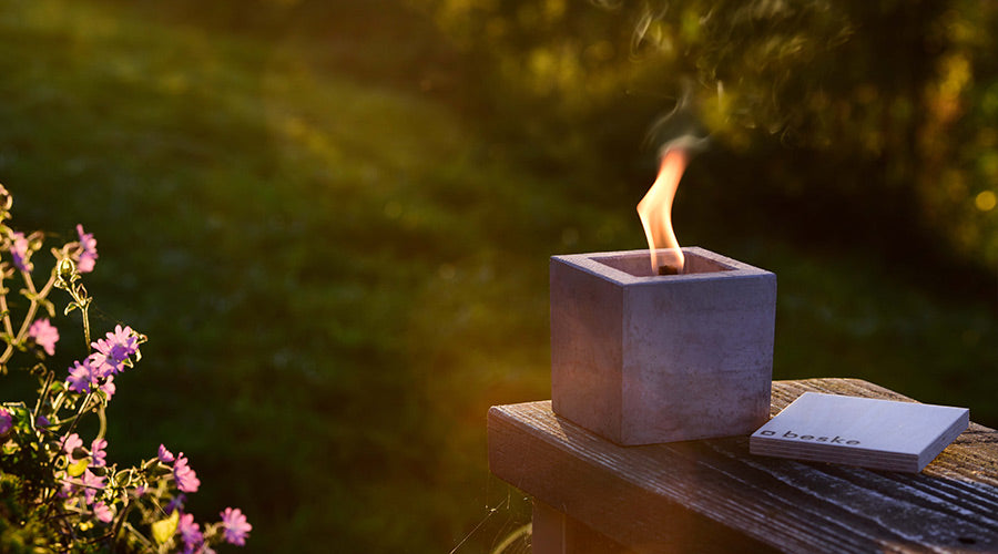 Betonfeuer brennt auf einem schmalen Holztisch in der sommerlichen Dämmerung, mit Löschbrett daneben und sanftem Sonnenlichteinfall, umgeben von einer idyllischen Atmosphäre.