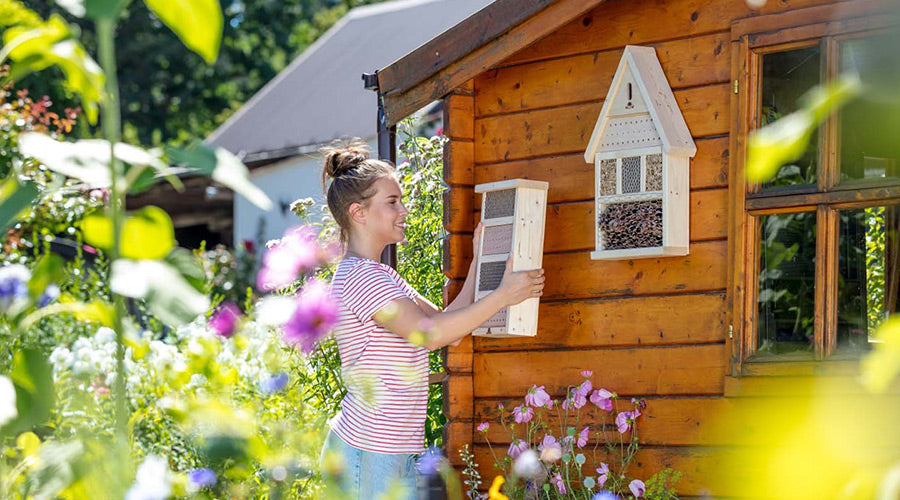 Junge Frau befestigt lächelnd ein Bienenhotel an einer Holzwand, neben einem Insektenhotel, umgeben von grüner Gartenlandschaft