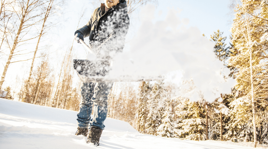 Mann im Schnee, kaum erkennbar, wirft Schnee in Richtung Kamera. Fokus auf fliegendem Schnee von der Schneeschaufel, die er mit beiden Händen hält, vor einem Waldhintergrund.