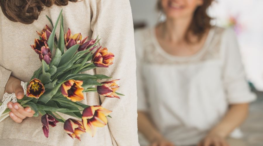 Kind hält einen Tulpenstrauß hinter dem Rücken als Überraschung zum Muttertag, im Hintergrund die Mutter.