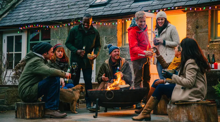 Gruppe von etwa 10 Personen um eine Feuerschale, trinken Bier und halten sich warm, dick gekleidet. Im Hintergrund ein Haus mit Lichterkette, vermutlich Herbstgeburtstag.