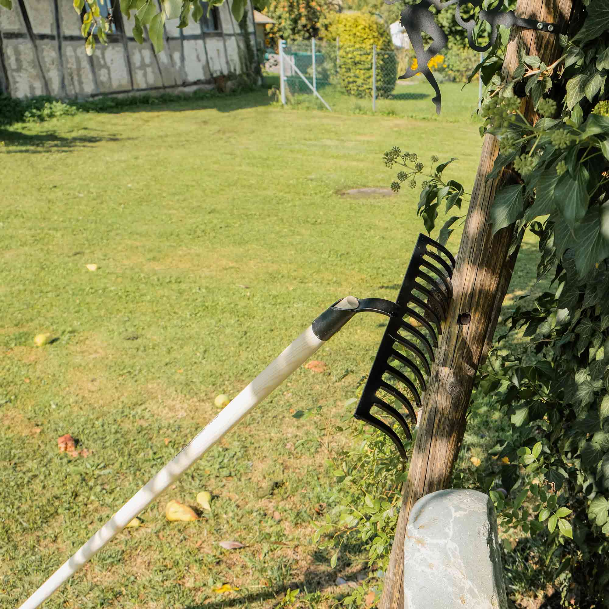 Gartenrechen mit gebogenem Stahlkopf und Holzstiel an einem Holzbalken im Garten gelehnt