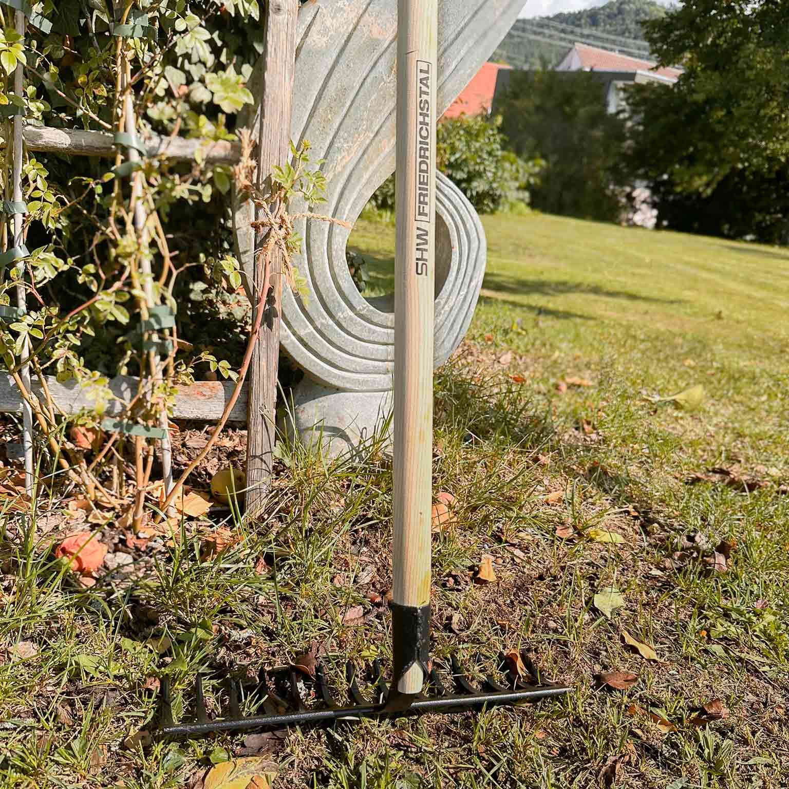 Rechen mit stabilem Holzstiel und schwarzem Stahlkopf stehend auf Rasenfläche neben Skulptur