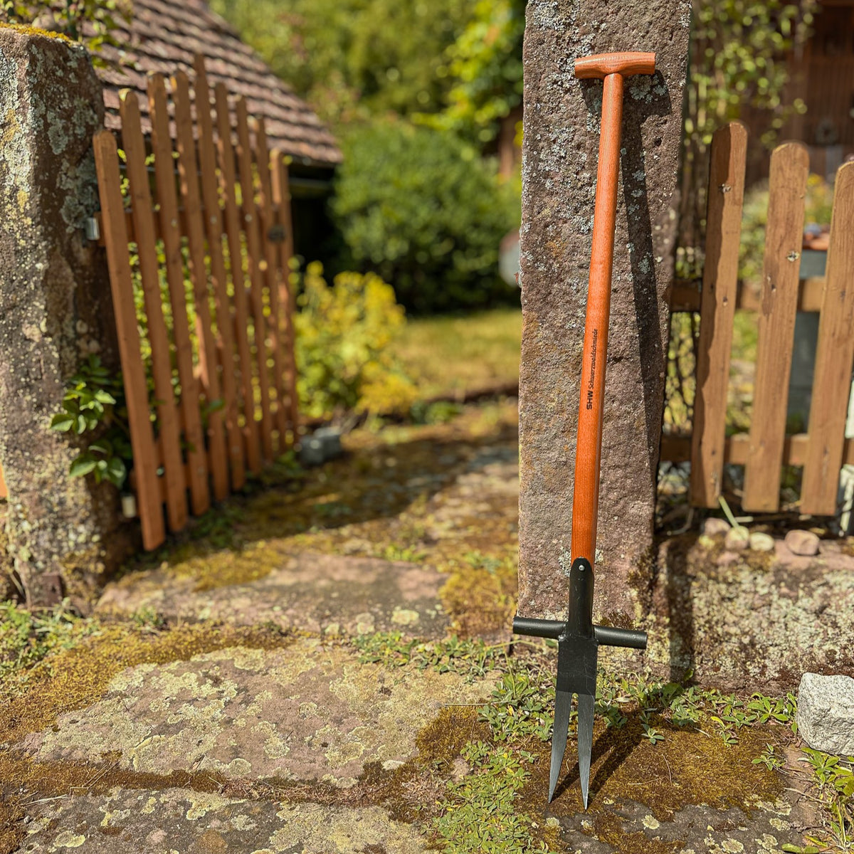 SHW Ampferausstecher mit zwei langen, spitzen Stahlzinken, seitlichen Tritthilfen und stabilem Holzstiel mit T-Griff, aufgestellt an einem Natursteinpfosten im Garten.