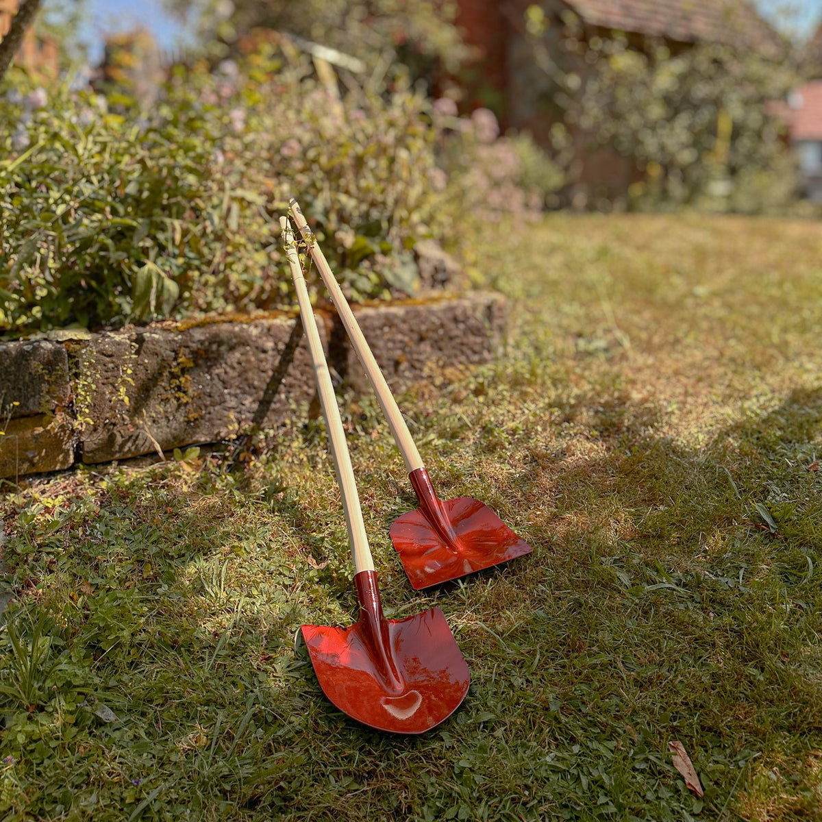 Zwei rote SHW Frankfurter Schaufeln mit Aluminiumblatt und langen Holzstielen stehen auf einer Rasenfläche im Garten. Unterschiedliche Schaufelblattformen sind gut erkennbar.