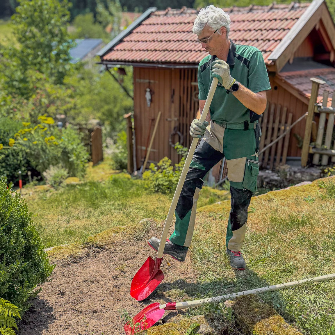 Gärtner arbeitet mit einer roten SHW Frankfurter Schaufel mit Aluminiumblatt und langem Holzstiel an einem Gartenbeet. Die Schaufel wird zum Aufnehmen und Bewegen von Erde eingesetzt.