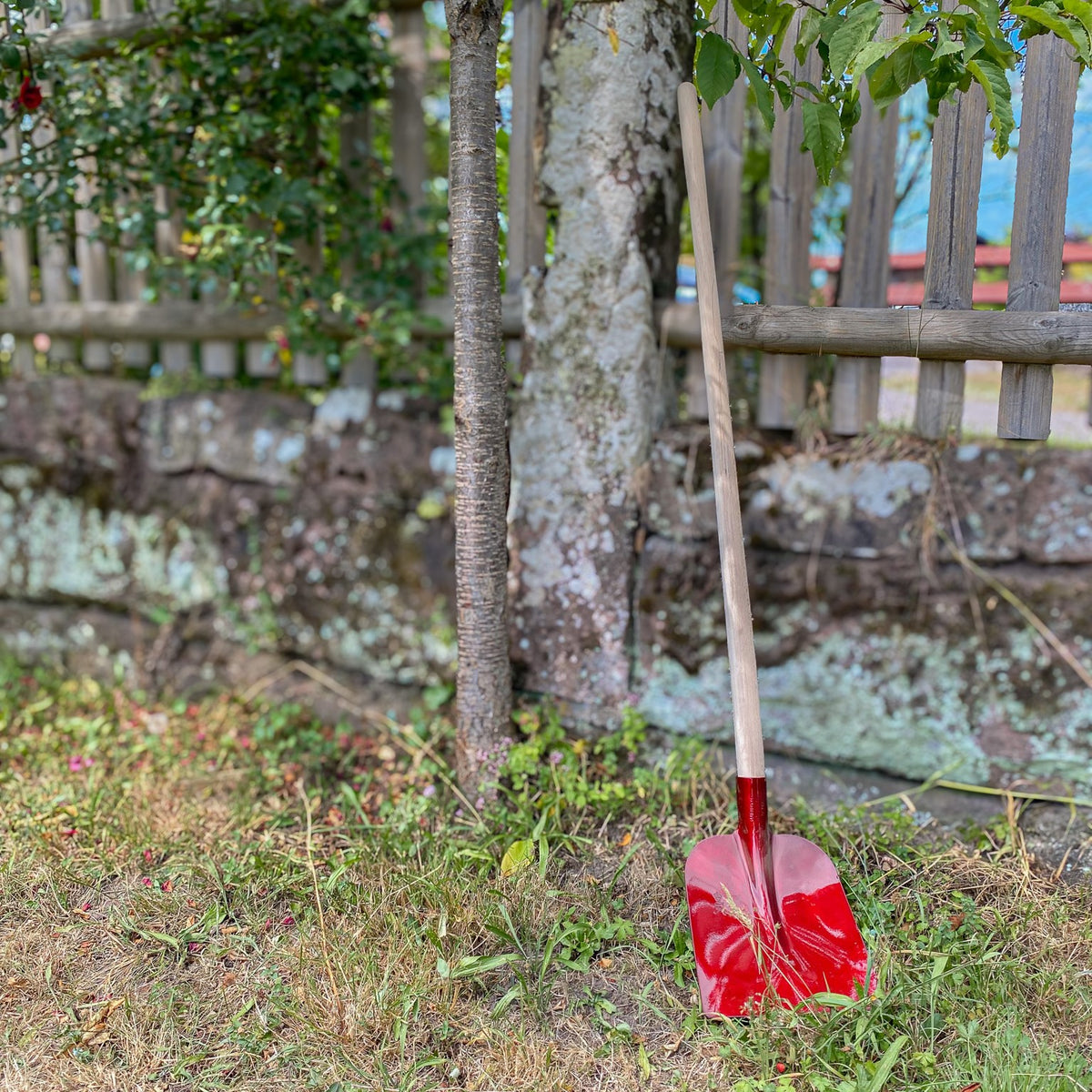 Rote SHW Holsteiner Schaufel aus Aluminium mit langem Holzstiel, aufgestellt auf einer Rasenfläche vor einem Holzzaun und einer Natursteinmauer im Garten.