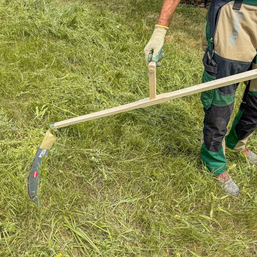 Gärtner hält die SHW Sense Friedrichstal mit Holzworb und gebogenem Sensenblatt auf einer Wiese mit frisch geschnittenem Gras. Traditionelle Handsense zur manuellen Wiesenmahd.